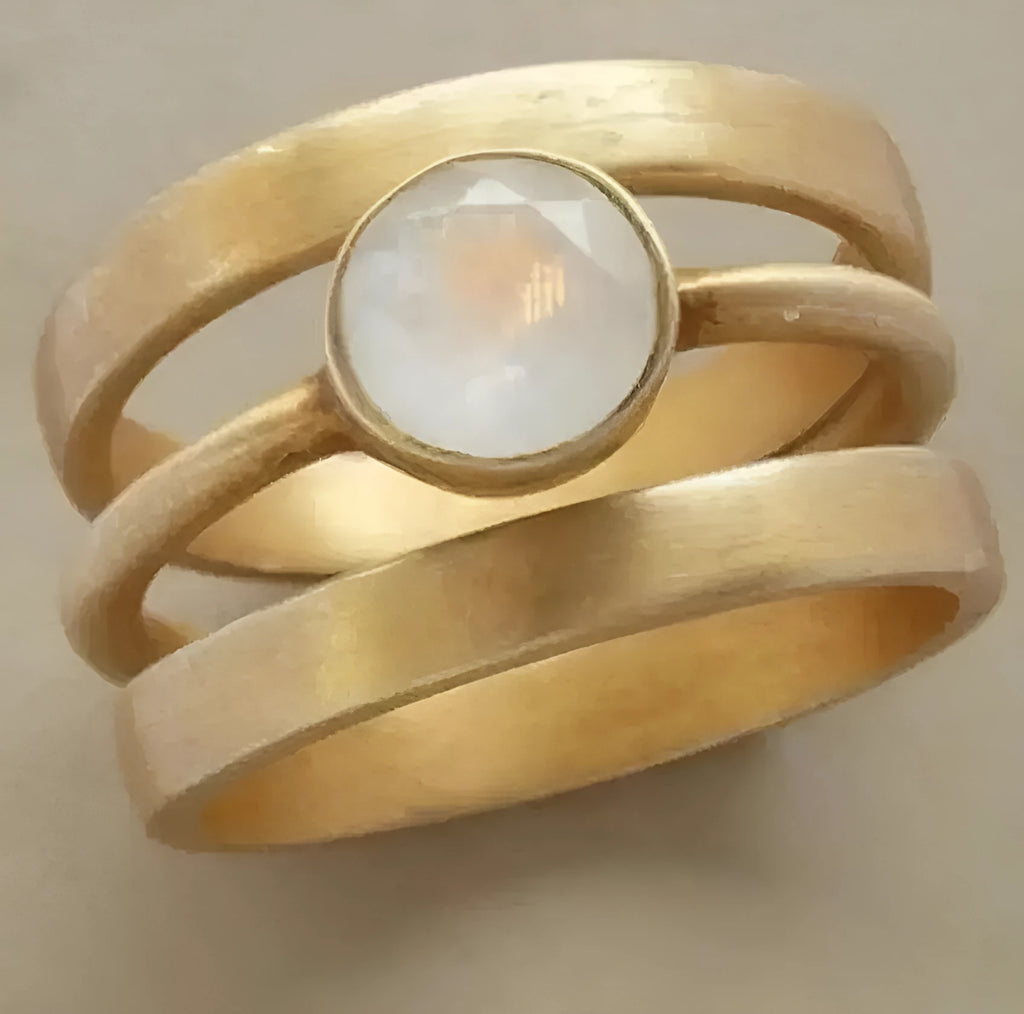 A gold geometric ring with linear design and a micro-inlaid white chalcedony stone on a grey background.
