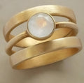A gold geometric ring with linear design and a micro-inlaid white chalcedony stone on a grey background.