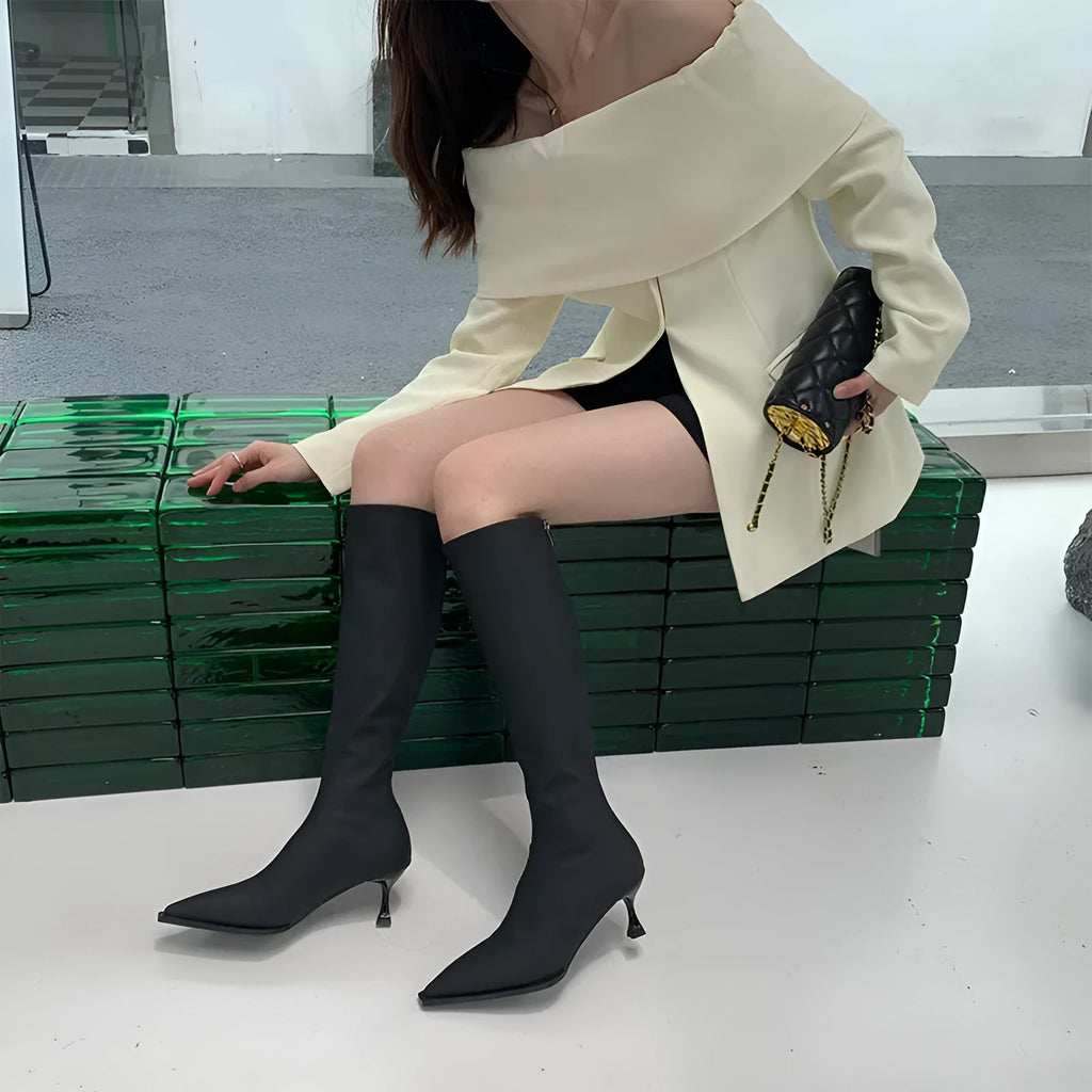 Black pointed toe knee-high boots with a stiletto heel and back zipper, shown on a model