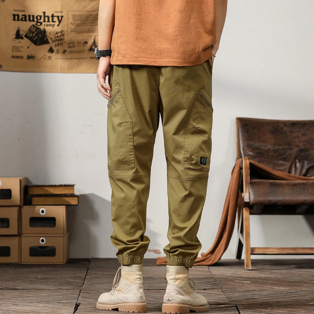 Men's khaki cotton tapered harem pants with ankle bands and a zip fly, laid flat.