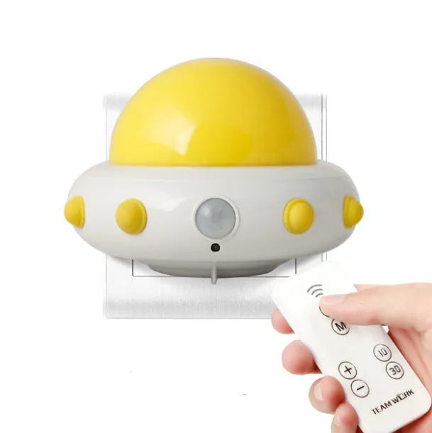 Remote control UFO shaped bedside night light in blue, pink, or white on a bedside table.