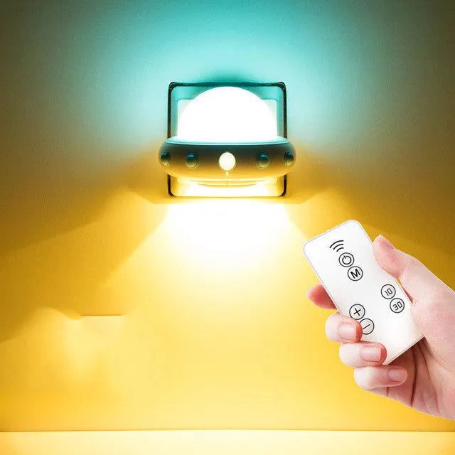 Remote control UFO shaped bedside night light in blue, pink, or white on a bedside table.