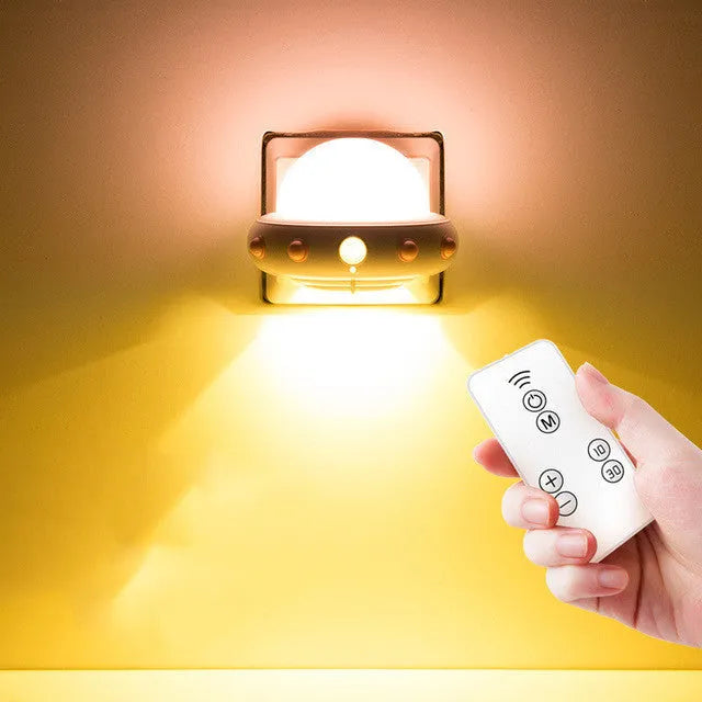Remote control UFO shaped bedside night light in blue, pink, or white on a bedside table.