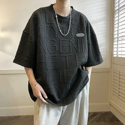 Man wearing a grey loose-fit T-shirt with a retro-style 3D raised letter print across the chest.