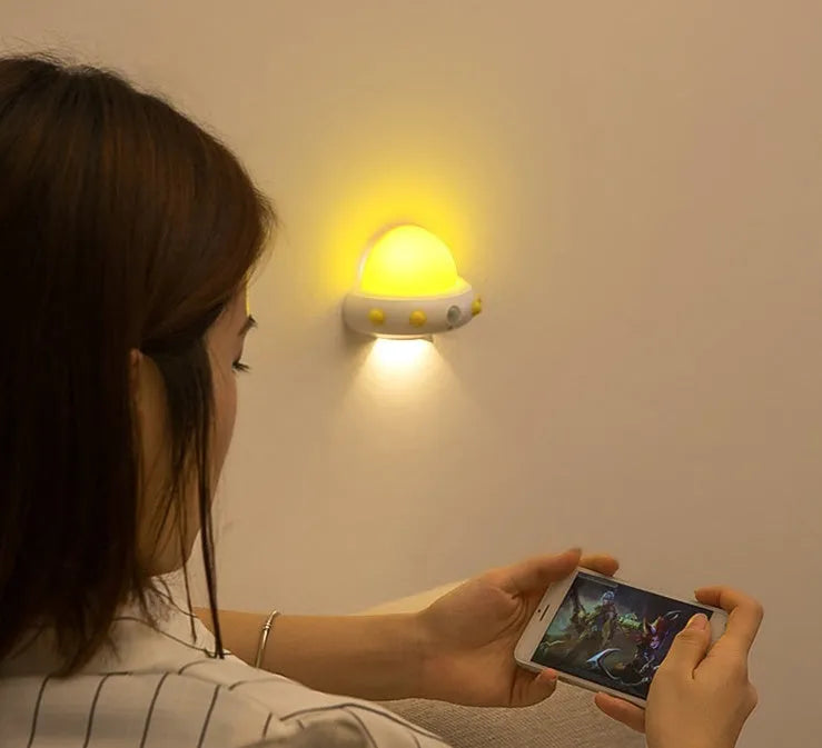 Remote control UFO shaped bedside night light in blue, pink, or white on a bedside table.
