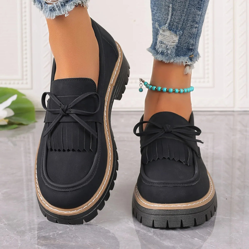 Black women's loafers with a tassel detail and chunky rubber sole on a wooden floor.