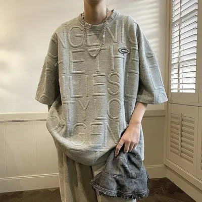 Man wearing a grey loose-fit T-shirt with a retro-style 3D raised letter print across the chest.